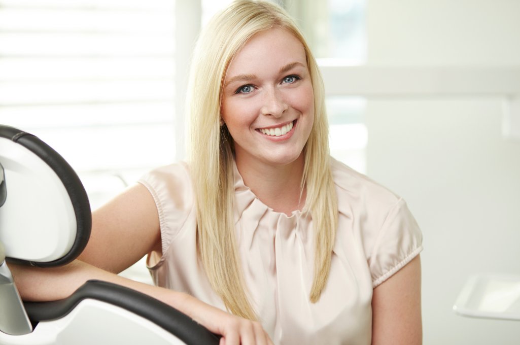 frau-laechelt-zahnarztstuhl Eine blonde Frau sitzt in einem Zahnarztstuhl und lächelt in die Kamera.